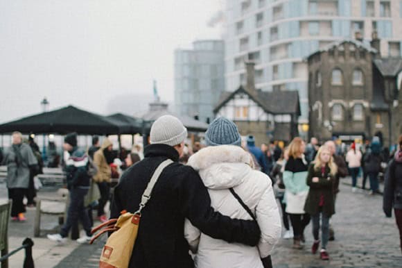 A couple, dressed in layers, including beanies and jackets, walking through a busy European street market.